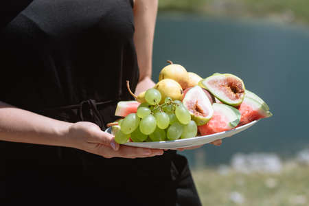 Young woman in a black dress holds a plate of fruit: pears, watermelon, grapes and figs at picnic in the mountains Montenegro.の写真素材
