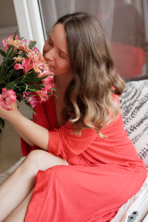 Young Blonde woman in a red robe with flowers against wide window background having lazy morningの写真素材