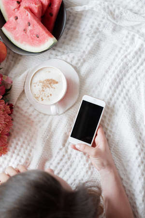 Top view of a Pretty blonde woman in a red robe using phone on the bed and drinks coffee against window backgroundの写真素材