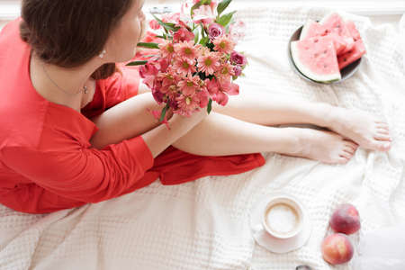 Beautiful Legs close-up in Bed, woman in a red robe  drinks coffee against wide window background and having lazy morningの写真素材