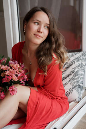 Young Blonde woman in a red robe with flowers against wide window background having lazy morningの写真素材