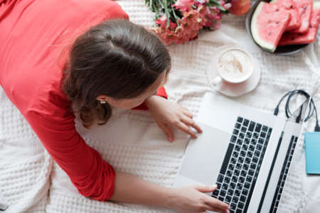 Top view of blonde woman in a red robe working on laptop on the bed and drinks coffee against window backgroundの写真素材