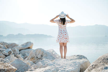 Back view of a young beautiful woman holding her white hat at the beach and looking to the horizon.の写真素材