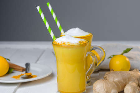 Turmeric latte or golden milk in glasses with ginger and lemons on a white tableの写真素材