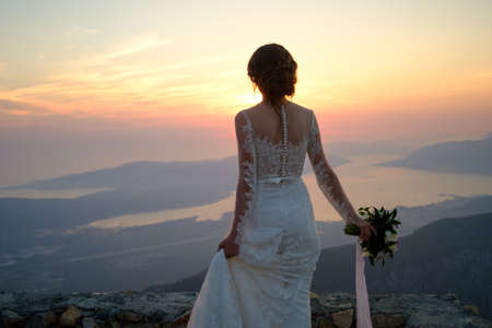 back of the bride in white dress standing on top of the mountain at sunsetの写真素材