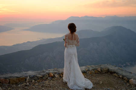 back of the bride in white dress standing on top of the mountain at sunsetの写真素材