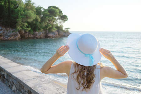 Back view of a young beautiful woman holding her straw hat at the beach and looking to the  sea horizon.の写真素材