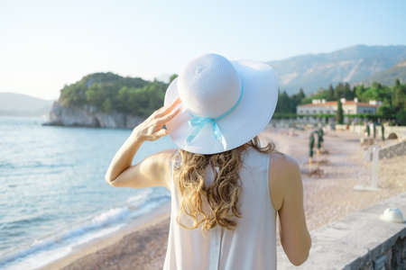 Back view of a young beautiful woman holding her straw hat at the beach and looking to the  sea horizon.の写真素材