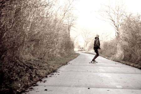 Autumn day with falling leaves from trees  A young blonde girl with longboard rolls down the asphalt road in a gray environment の写真素材