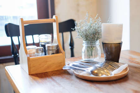 Beautiful houseplant in flowerpot, tissue in black box, garnish (pepper, toothpick, sauce) in wooden box and spoon/fork set on wooden table and black chair, for food background - interiors concept.の写真素材