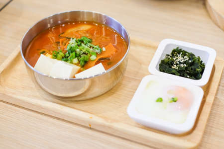 The homemade food set consists of spicy kimchi soup (bacon, slice pork, white tofu, spring onion & cabbage), soft boiled egg (onsen), green seaweed with white sesame serve on wooden tray.の写真素材