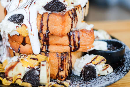 Close up triple toast bread topped with chocolate sauce, cereals, cookies, white cream, banana and vanilla ice cream serve on black dish for dessert background or texture.の写真素材