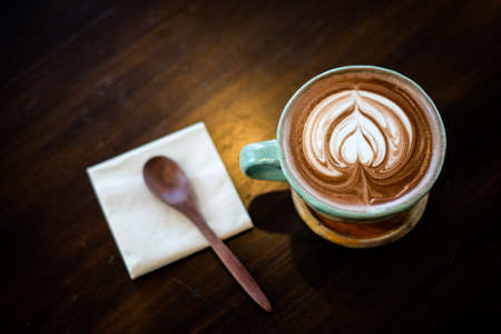 Homemade hot fresh cocoa with patterned white milk froth on surface in cup with wooden saucer (bottom plate), spoon and tissue serve on wooden table, for beverage background - healthy diet concept.の写真素材