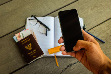 Man's hand using mobile phone (black screen) with Thailand passport, money, yellow ruler, pencil, notebook and glasses for contact business and prepare travel.の写真素材