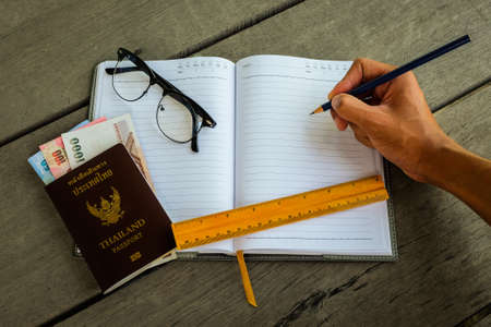 Thailand passport with money, yellow ruler, hand with pencil writing on notebook and glasses - concept prepare to travel.の写真素材