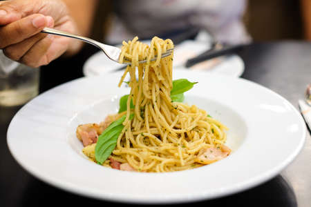 Woman's hand holding fork for eating spaghetti spicy bacon & ham with basil, tomato, black pepper, lettuce and paprika on white dish set on table for food background or texture.の写真素材