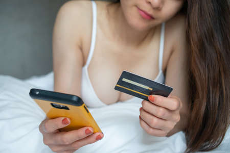 Cropped shot of sexy woman lying on bed and using her mobile phone for shopping online and bought...