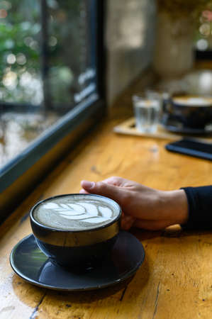 Cropped shot view of someone hand touching a cup of Goth Latte or Charcoal Latte on wooden table....