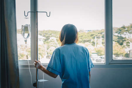 Back view of sickness patient woman standing nearly window in hospital ward, looking to outside and hope her health will be better. Conceptual of sickness woman feeling when admitted in the hospital.の写真素材