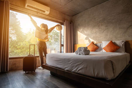 Portrait of tourist woman raised her hands and standing nearly window, looking to beautiful view with her luggage in hotel bedroom after check-in. Conceptual of travel and vacation.の写真素材
