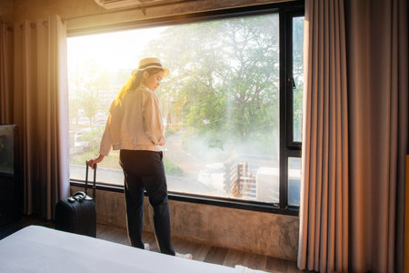 Portrait of tourist woman standing nearly window with her luggage in hotel bedroom after check-in. Conceptual of travel and vacation.の写真素材