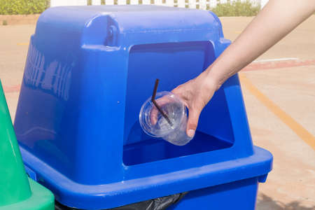 Close-up hand throwing empty plastic glass in the blue recycling bin. A container used to hold...