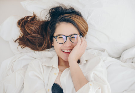 Portrait of young attractive Asian woman lying and relaxing on comfortable bed and looking to camera. Conceptual of woman lifestyle while waking up on bed.の写真素材