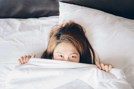 Close-up of cute lovely young Asian woman lying and hiding under white blanket in her bedroom.の写真素材