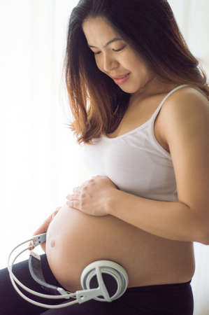 Young Asian pregnant woman sitting and relax in her bedroom and using headphone on her belly for her unborn child listens to classic music.の写真素材