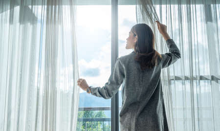 Rear view of happiness Asian woman open curtains for looking beautiful view outside her room.