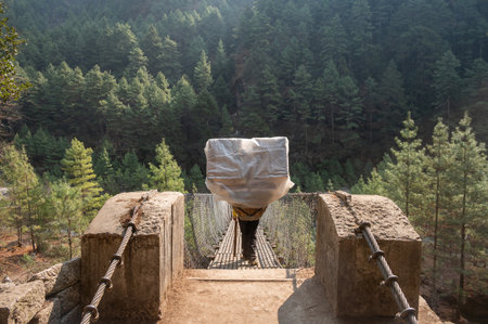 Rear view of Nepali porter carrying stuff while walking on the suspension bridge for crossing river...
