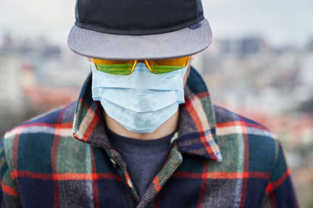 Fashion styled distressed young man in medical mask and goggles with blurred city view backgroundの写真素材