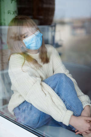 Young attractive woman in white sweater and jeans with surgical mask looking out of home window,...