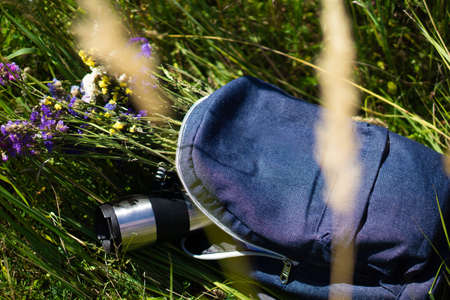 Blue jeans school backpack lying on green grass with travel mug and meadow flowers inside it. Relaxation and summer hiking conceptの写真素材