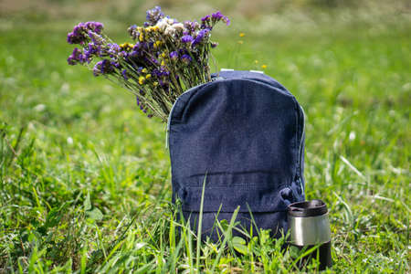 Relaxation and summer hiking concept. Blue jeans school backpack standing on green grass with travel mug near it and beautiful meadow flowers inside itの写真素材