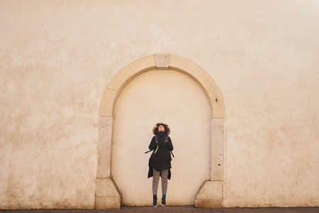 Young brunette tourist woman with curly hair and black coat jumping with big wall background and looking up. Lifestyle and traveling concept.の写真素材