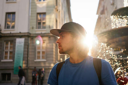 Portrait of attractive young bearded smiling tourist man in baseball hat and t-shirt looking left side with old city background and sunset with fountain on background. Lifestyle and traveling conceptの写真素材