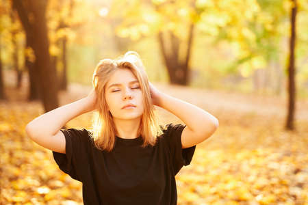 Young blonde student girl in black t-shirt posing in autumn park with closed eyes and hands on a...