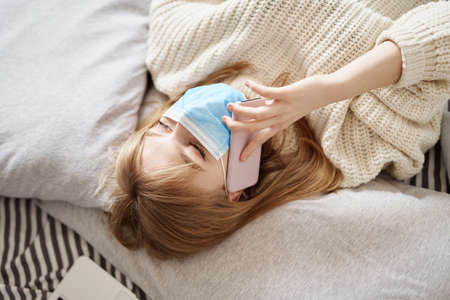 Isolation at home: young attractive teen in medical mask wearing white sweater lying on a bed on quarantine talking on a cell phoneの写真素材