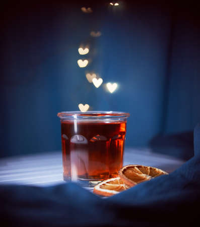 Glass of warm tea with orange slices on the blue background with blurred lights in form of heart in...