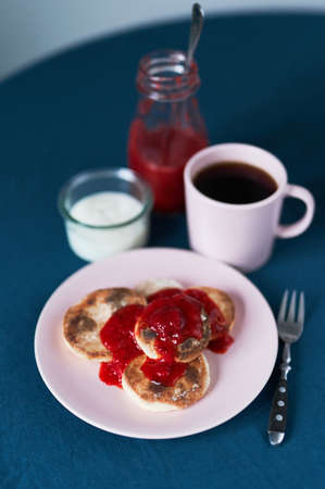 Fresh cheese pancakes with cream and strawberries jam with cup of black coffee on a blue background. Tasty breakfast concept.の写真素材