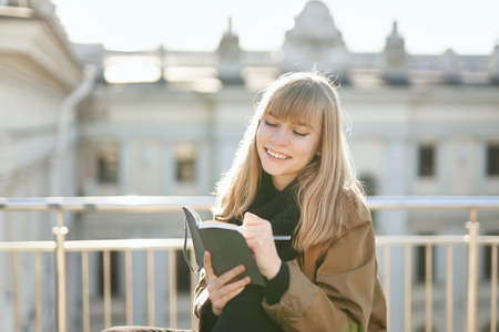 Blonde student girl smiling and writing in the notepad during outdoor working. European city concept