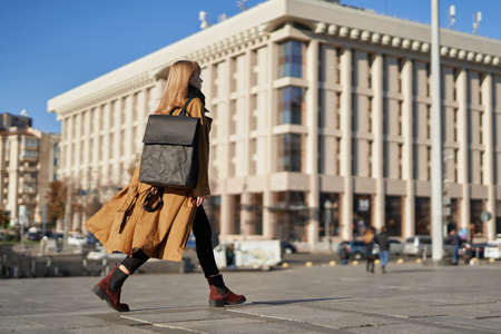 Blonde girl in orange trench walking with black ecological paper backpack in European cityの写真素材