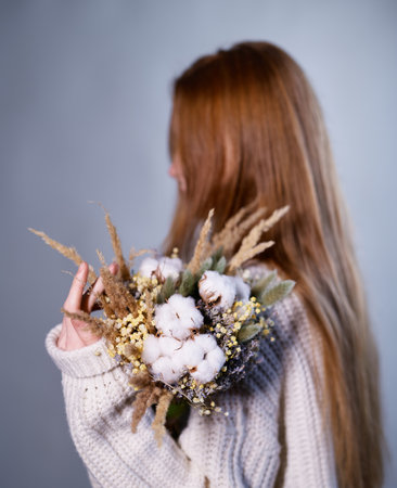 Attractive girl in white sweater holding bouquet of beautiful dried flowers against a gray wall background. A faceless girl holds a bouquet. Flower delivery incognitoの写真素材