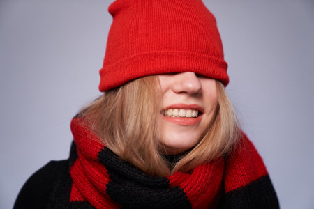 Pretty blonde smiling girl with red and black scarf and hat over eyes isolated on gray background. Winter, warm clothes conceptの写真素材