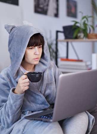 Business woman in bathrobe drinks morning coffee and surfing internet on laptop. Cheerful girl writing email on computer or working in home office. Remote studying or working conceptの写真素材