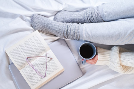 Aesthetic photo of woman on bed in sweater, warn socks, book, glasses and cup of coffee in hands, top view point. Lazy weekend conceptの写真素材