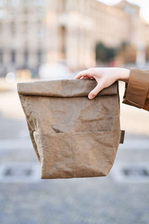 Hand holding reusable paper lunch bag on the background of the city view. Eco lunch box. Lunchtime conceptの写真素材