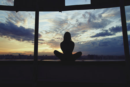 Silhouette of young woman practicing yoga in urban surroundings. Practicing yoga outdoors at sunset with a big city at the background. Healthy, harmony and meditation conceptの写真素材