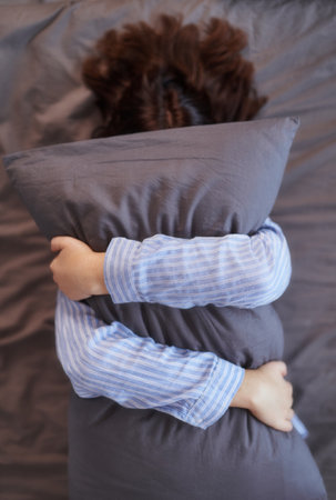 Too lazy to get out of bed concept. Young brunette woman in pajama lying on a bed and covers her face hugging a gray pillowの写真素材
